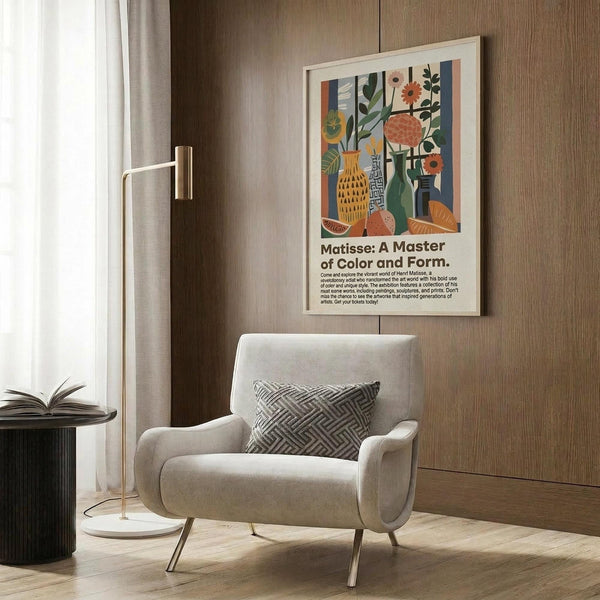 May include: Modern living room setting with beige upholstered armchair featuring geometric patterned pillow, gold floor lamp, dark round side table with open book, and wood-paneled wall. Framed poster displays colorful still life artwork with vases, plants, and fruit in vibrant oranges, greens, and blues alongside exhibition text about Matisse's artistic style and color mastery.