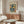 May include: Matisse art poster featuring abstract blue figure in cut-out style with colorful geometric shapes in yellow, orange, and red, displayed in thin brown frame above mid-century walnut credenza. Modern living room setting with cream textured chair, glass oval coffee table, and white table lamp. Minimalist Scandinavian interior design with neutral walls and natural lighting.