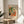 May include: Framed Matisse-style poster print displayed on light wood round side table next to beige upholstered chair. Artwork features colorful interior scene with orange flowers in black vase, green palm plants, leopard print pillow on coral chair, and geometric patterns in warm tones. Natural sunlight creates soft shadows on white wall. Real plant leaves visible in corner of minimalist room setting.