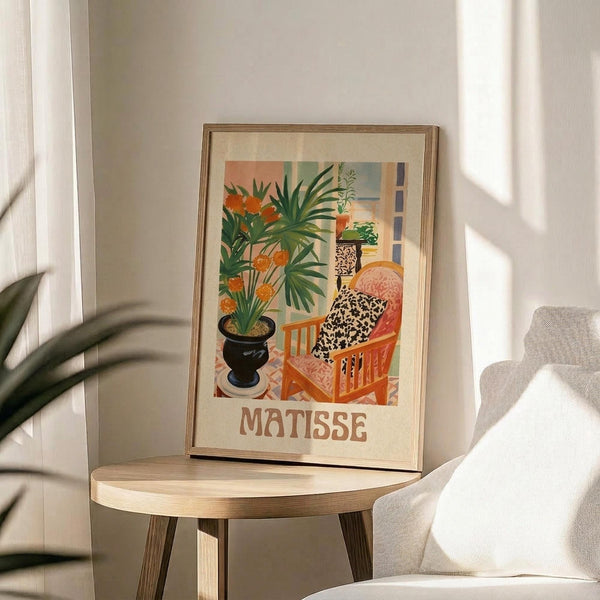 May include: Framed Matisse-style poster print displayed on light wood round side table next to beige upholstered chair. Artwork features colorful interior scene with orange flowers in black vase, green palm plants, leopard print pillow on coral chair, and geometric patterns in warm tones. Natural sunlight creates soft shadows on white wall. Real plant leaves visible in corner of minimalist room setting.