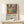 May include: Framed poster displaying colorful still life artwork with vases, flowers, and fruit in vibrant oranges, greens, and blues, positioned on light wood surface with small wrapped gift box. Exhibition-style poster features descriptive text below the artwork. Soft natural lighting creates gentle shadows on neutral wall background.