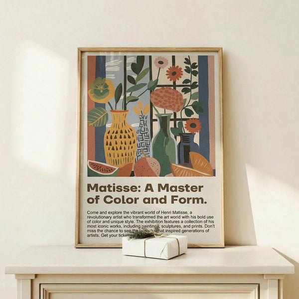 May include: Framed poster displaying colorful still life artwork with vases, flowers, and fruit in vibrant oranges, greens, and blues, positioned on light wood surface with small wrapped gift box. Exhibition-style poster features descriptive text below the artwork. Soft natural lighting creates gentle shadows on neutral wall background.