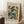 May include: Framed art poster displaying abstract cut-out style figures in teal, orange, yellow and black colors against cream background, propped against white paneled wall on textured white rug. Natural wood frame surrounds the modernist artwork. White coffee cup on wooden block sits nearby next to beige fabric chair, creating cozy room setting with soft natural lighting.