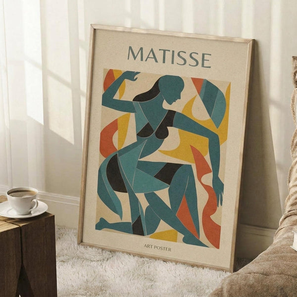 May include: Framed art poster displaying abstract cut-out style figures in teal, orange, yellow and black colors against cream background, propped against white paneled wall on textured white rug. Natural wood frame surrounds the modernist artwork. White coffee cup on wooden block sits nearby next to beige fabric chair, creating cozy room setting with soft natural lighting.