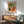 May include: Framed Matisse-style poster featuring woman in leopard print dress sitting in orange chair beside large potted plant with orange flowers, displayed above wooden sideboard with white lamp in modern living room setting with sage green armchair and natural lighting casting shadows on white wall.