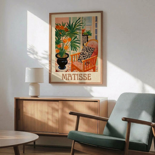 May include: Framed Matisse-style poster featuring woman in leopard print dress sitting in orange chair beside large potted plant with orange flowers, displayed above wooden sideboard with white lamp in modern living room setting with sage green armchair and natural lighting casting shadows on white wall.