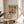 May include: Framed botanical art print with colorful abstract flowers and leaves in pink, orange, green, and blue tones, displayed on round wooden side table next to beige upholstered chair. Sunlight casts geometric shadows on white wall behind the artwork. Plant leaves visible in corner of bright, minimalist living room setting.