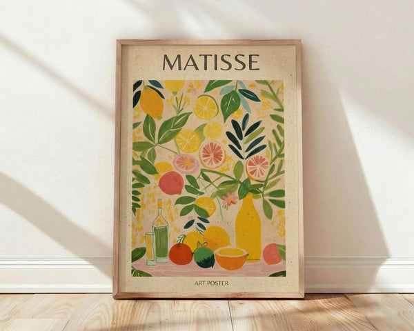 May include: Framed art poster in natural wood frame leaning against white wall on light wooden floor. Artwork features colorful still life with citrus fruits, leafy branches, bottles and bowls in Matisse-inspired style using yellows, greens, oranges and pinks on cream background. Text reads "MATISSE" at top and "ART POSTER" at bottom. Soft natural lighting creates gentle shadows on wall.
