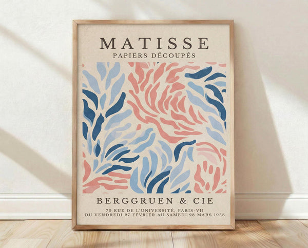 May include: Framed poster print in natural wood frame leaning against white textured wall on light wooden floor. Features abstract cut-out style artwork with flowing organic shapes in coral pink and various shades of blue on cream background. French exhibition text at top and bottom. Soft natural lighting creates gentle shadows on wall.