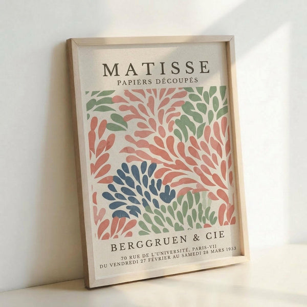 May include: Framed vintage Matisse exhibition poster featuring colorful paper cut-out leaf patterns in coral pink, sage green, and navy blue on cream background. Light wood frame displayed against white wall, showing gallery text for Berggruen & Cie Paris exhibition from 1953.