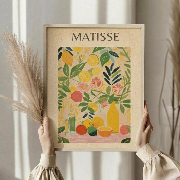 May include: Person in beige ruffled sleeves holding framed botanical poster print featuring colorful citrus fruits, leaves, and bottles in Matisse-inspired cut-out style with yellows, greens, pinks, and oranges on cream background in light wooden frame, with dried pampas grass visible beside neutral curtains.
