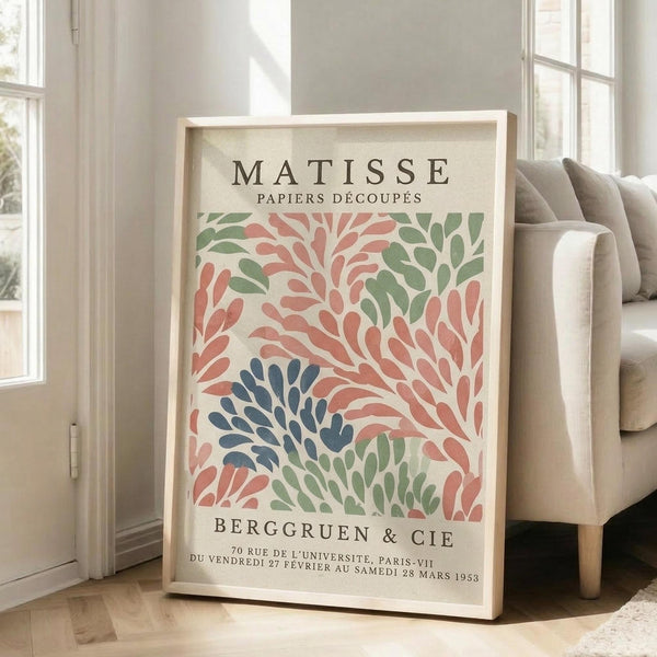 May include: Framed vintage-style exhibition poster featuring abstract cut-out leaf shapes in coral pink, sage green, and navy blue on cream background. Light wood frame leans against white wall next to beige upholstered sofa in bright, airy living room with large windows and natural lighting.