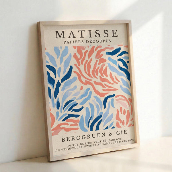 May include: Framed vintage Matisse exhibition poster leaning against white wall. Features abstract coral and blue organic shapes resembling seaweed or leaves on cream background. Text reads "MATISSE PAPIERS DÉCOUPÉS" at top and "BERGGRUEN & CIE" with Paris exhibition details at bottom. Natural wood frame with visible grain texture.