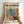 May include: Hands holding a framed poster with colorful botanical leaf cutouts in Matisse style, featuring vibrant pink, teal, orange, yellow and black tropical foliage shapes on cream background. Light wood frame against white wall, natural lighting creates soft shadows.