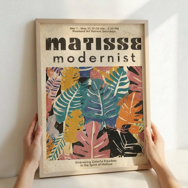 May include: Hands holding a framed poster with colorful botanical leaf cutouts in Matisse style, featuring vibrant pink, teal, orange, yellow and black tropical foliage shapes on cream background. Light wood frame against white wall, natural lighting creates soft shadows.