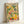 May include: Framed art poster with Matisse-inspired design featuring colorful citrus fruits, green foliage, and bottles arranged in still life composition. Bright yellows, oranges, greens and coral colors on cream background. Light wood frame displayed against white wall, showing natural lighting and shadows.