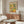 May include: Colorful Matisse-style poster featuring vibrant citrus fruits, lemons, oranges, and green foliage in cut-out art style, displayed in thin wooden frame above mid-century walnut credenza with white table lamp, cream upholstered chair, and glass oval coffee table in bright modern living room with white walls and natural lighting