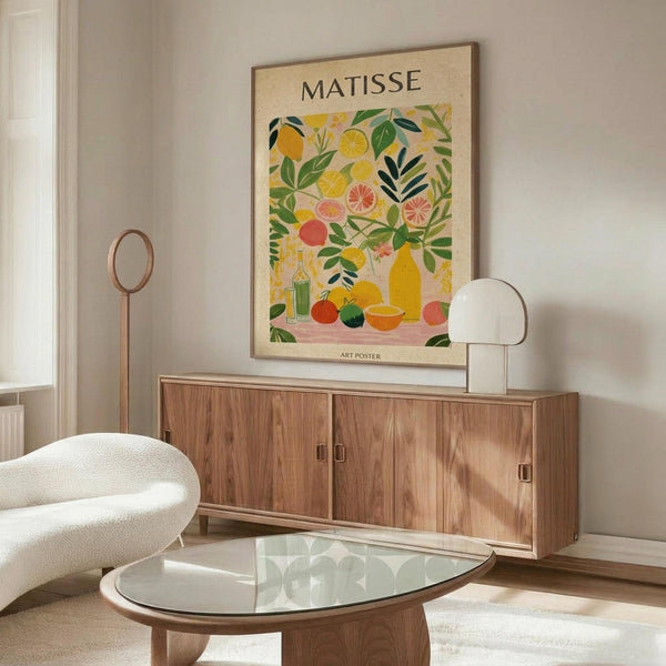 May include: Colorful Matisse-style poster featuring vibrant citrus fruits, lemons, oranges, and green foliage in cut-out art style, displayed in thin wooden frame above mid-century walnut credenza with white table lamp, cream upholstered chair, and glass oval coffee table in bright modern living room with white walls and natural lighting