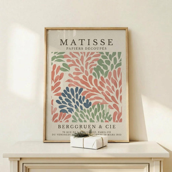 May include: Framed art print featuring Matisse-style cut paper botanical design with coral pink, sage green, and navy blue leaf shapes on cream background. Displayed on light wood console table with small white gift box and evergreen sprig against neutral wall.