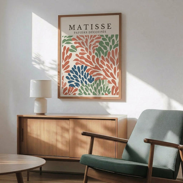 May include: Framed Matisse cut-out style art print with organic leaf and coral forms in coral, green, and navy blue colors, displayed above a mid-century modern wooden credenza with ceramic table lamp, next to a sage green upholstered armchair with wooden arms in a sunlit room with white walls