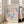 May include: Framed exhibition poster with abstract coral and seaweed-like shapes in coral pink and navy blue tones, leaning against white wall next to beige upholstered sofa in bright sunlit room with large windows and wooden floors.