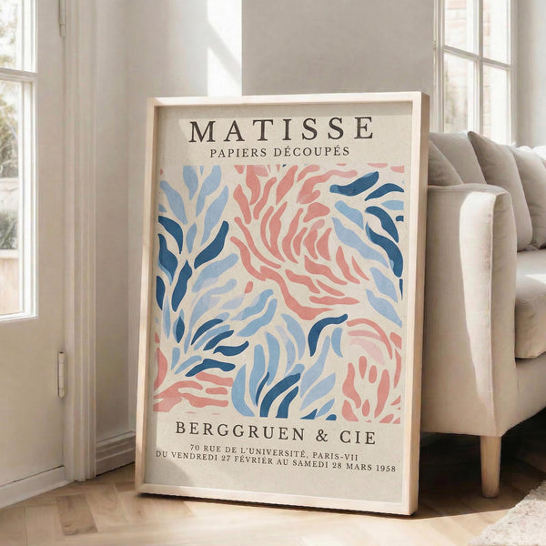 May include: Framed exhibition poster with abstract coral and seaweed-like shapes in coral pink and navy blue tones, leaning against white wall next to beige upholstered sofa in bright sunlit room with large windows and wooden floors.