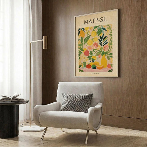May include: Colorful Matisse-inspired poster with vibrant fruits, citrus slices, and leafy botanicals in yellows, greens, and coral tones, displayed in light wood frame above beige upholstered chair with geometric pillow. Modern minimalist room setting features brass floor lamp, dark round side table with open book, wood paneled wall, and white curtains.
