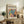 May include: Framed poster with colorful tropical leaves in pink, teal, orange, and yellow tones displayed on rustic wooden sideboard beside fern plant, ceramic vases, and stacked books in neutral-toned living room setting