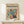 May include: Framed art print featuring colorful botanical leaf patterns in teal, coral, yellow and black, displayed on light wood surface next to small white wrapped gift with twine and greenery sprig against cream wall