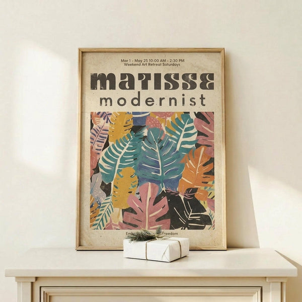 May include: Framed art print featuring colorful botanical leaf patterns in teal, coral, yellow and black, displayed on light wood surface next to small white wrapped gift with twine and greenery sprig against cream wall
