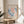 May include: A framed vintage Matisse exhibition poster featuring abstract coral and blue leaf-like shapes in his signature cut-out style. The cream-colored print sits on a round wooden stool next to a beige upholstered chair, with soft natural lighting creating shadows on white walls and a plant visible in the corner.