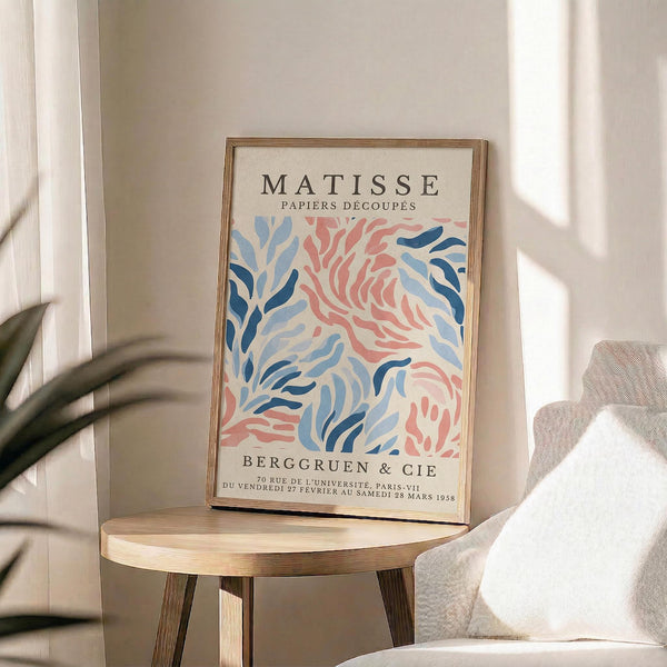 May include: A framed vintage Matisse exhibition poster featuring abstract coral and blue leaf-like shapes in his signature cut-out style. The cream-colored print sits on a round wooden stool next to a beige upholstered chair, with soft natural lighting creating shadows on white walls and a plant visible in the corner.