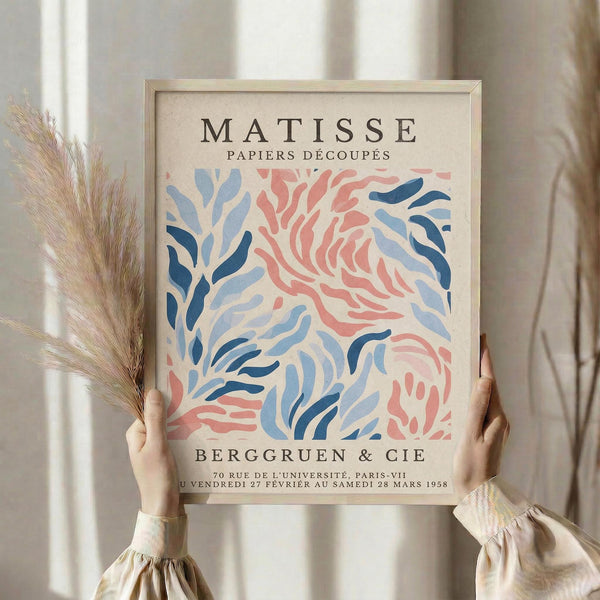 May include: Person holding framed vintage art exhibition poster featuring Matisse's papiers découpés cut-out style in coral pink and blue organic shapes against cream background. Light wood frame, dried pampas grass in other hand, neutral curtained interior setting with soft natural lighting.