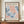 May include: Framed vintage art exhibition poster with abstract organic shapes in coral pink and blue tones resembling flowing seaweed or coral forms against cream background. Features French text including gallery name and 1958 exhibition dates. Displayed on warm wood sideboard next to small white ceramic vase and stacked books in neutral-toned room with white paneled walls.