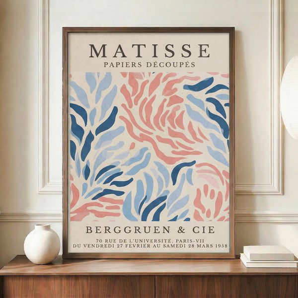 May include: Framed vintage art exhibition poster with abstract organic shapes in coral pink and blue tones resembling flowing seaweed or coral forms against cream background. Features French text including gallery name and 1958 exhibition dates. Displayed on warm wood sideboard next to small white ceramic vase and stacked books in neutral-toned room with white paneled walls.