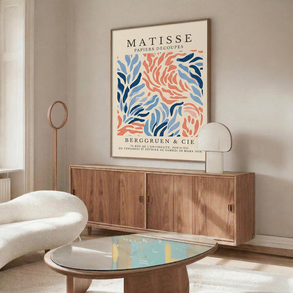 a living room with a large abstract art poster of the Matisse style on the wall above a wooden cabinet. The poster has a blue and orange color scheme with a leaf-like pattern. The room also includes a white sofa, a glass coffee table, and a lamp.