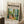 May include: Framed art poster featuring a colorful modernist-style interior scene with lush green plants, a yellow chair, and bright windows. The cream-colored poster displays "matisse modernist" text and event details. Set against white paneled walls on a textured rug, with a coffee cup on a wooden block and cozy blanket visible in the scene.