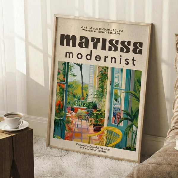 May include: Framed art poster featuring a colorful modernist-style interior scene with lush green plants, a yellow chair, and bright windows. The cream-colored poster displays "matisse modernist" text and event details. Set against white paneled walls on a textured rug, with a coffee cup on a wooden block and cozy blanket visible in the scene.
