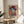 May include: Colorful art poster in wooden frame showing woman in hat rendered in vibrant blues, oranges, purples and geometric patterns with "MATISSE" text below, displayed on round wooden side table next to beige upholstered chair with plant leaves visible in soft natural lighting