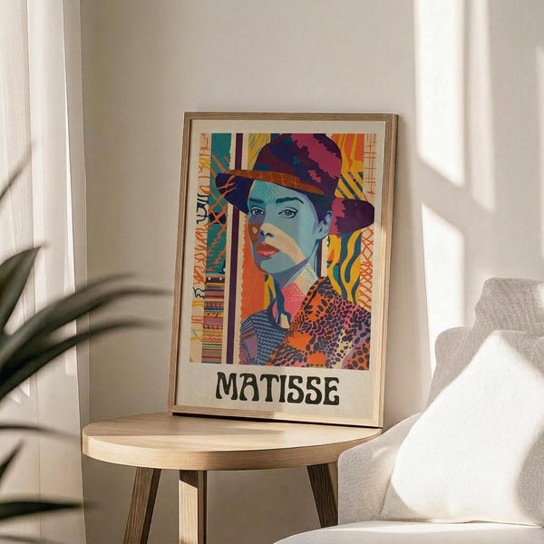 May include: Colorful art poster in wooden frame showing woman in hat rendered in vibrant blues, oranges, purples and geometric patterns with "MATISSE" text below, displayed on round wooden side table next to beige upholstered chair with plant leaves visible in soft natural lighting