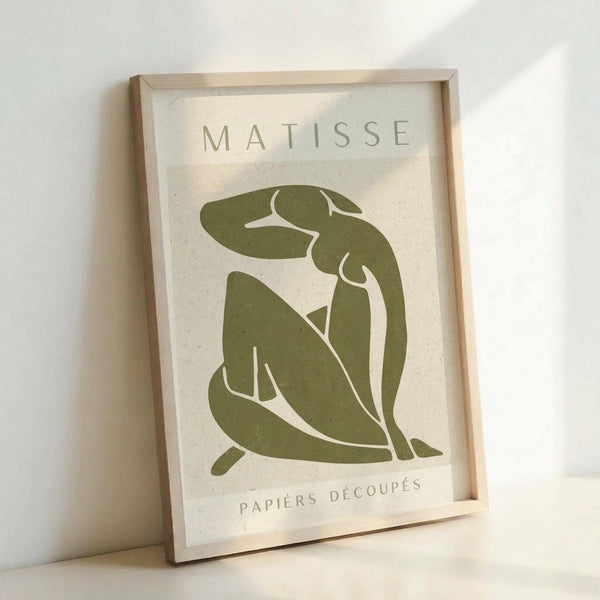 A framed art print featuring a sage green abstract figure in Matisse's cut-out style against a cream textured background. The minimalist design shows a seated nude form created from flowing organic shapes. "MATISSE" appears at the top and "PAPIERS DÉCOUPÉS" at the bottom in simple lettering. The print is displayed in a light wooden frame, leaning against a white wall.