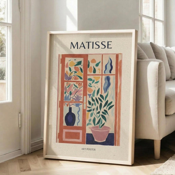 May include: Framed Matisse-inspired art poster leaning against white wall next to beige sofa. Poster features colorful cut-out style illustration of window with geometric panels showing plants, vases, and organic shapes in coral, blue, green, and pink tones. Light wood frame on hardwood floor in bright, minimalist living room with natural sunlight.