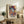 May include: Colorful portrait artwork in wooden frame showing woman with turban in vibrant blues, oranges, and purples with geometric patterns. Frame leans against white wall on rustic wood sideboard next to fern plant, books, and ceramic vase in warmly lit living room setting with beige armchair visible.