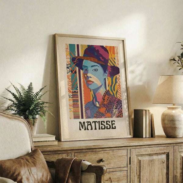 May include: Colorful portrait artwork in wooden frame showing woman with turban in vibrant blues, oranges, and purples with geometric patterns. Frame leans against white wall on rustic wood sideboard next to fern plant, books, and ceramic vase in warmly lit living room setting with beige armchair visible.