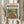May include: Person in cream sweater holding framed poster featuring colorful illustration of goldfish in glass bowl with orange and yellow flowers, styled in Matisse-inspired modernist art. Frame has light wood finish against neutral wall with dried pampas grass visible to left side.