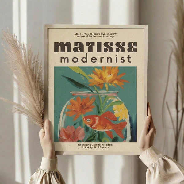 May include: Person in cream sweater holding framed poster featuring colorful illustration of goldfish in glass bowl with orange and yellow flowers, styled in Matisse-inspired modernist art. Frame has light wood finish against neutral wall with dried pampas grass visible to left side.
