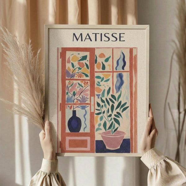 May include: Person in cream sweater holding framed Matisse-inspired art print featuring colorful botanical illustrations arranged in window pane layout. Artwork shows plants, leaves, and vase in warm orange, blue, green, and pink tones against cream background. Dried wheat stems visible beside figure with soft beige curtains in background.