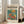 May include: Framed vintage-style art poster featuring a goldfish swimming in a clear glass bowl with colorful flowers - orange and pink blooms with green leaves against a teal background. The cream-colored poster has "matisse modernist" text and exhibition details. Light wood frame leans against white wall beside beige sofa in bright, naturally-lit living room with white window frames.