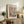 May include: Framed Matisse art poster with colorful cut-out style botanical illustrations in window panes, featuring plants, vase, and organic shapes in pink, blue, green, and orange. Displayed on rustic wooden sideboard alongside fern plant, ceramic vase, books, and white upholstered furniture in neutral-toned living room.