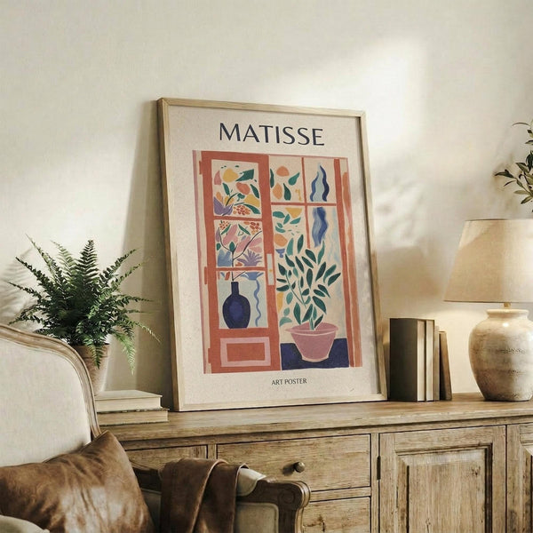 May include: Framed Matisse art poster with colorful cut-out style botanical illustrations in window panes, featuring plants, vase, and organic shapes in pink, blue, green, and orange. Displayed on rustic wooden sideboard alongside fern plant, ceramic vase, books, and white upholstered furniture in neutral-toned living room.
